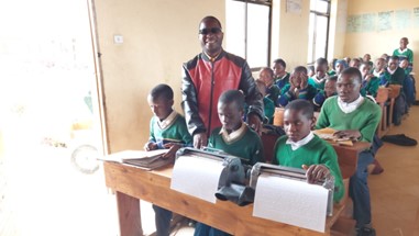 photo a Teacher Lazaro standing in a classroom full of seated students, beside three students reading and writing braille created on Perkins machines.