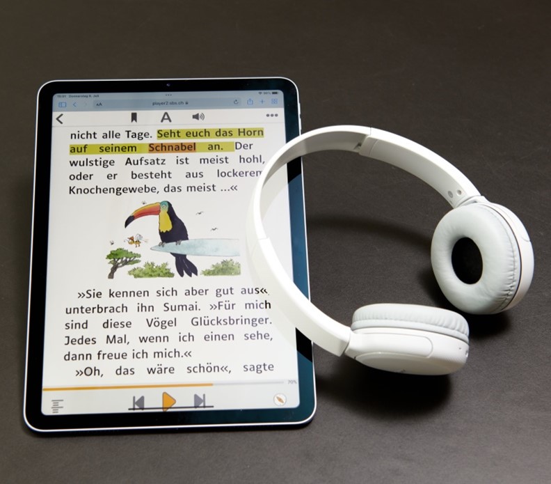 This image shows a tablet and a pair of headphones. The tablet is playing a Text-Audiobook using SBS online player. On screen you see the text with the highlighted words and a colourful drawing of a toucan bird.