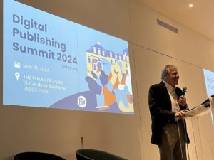 Photo of George Kerscher standing at a podium with a microphone, behind him a large Digital Publishing Summit 2024 slide is projected