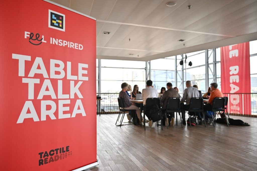 Photo of people gathered around a table in the "Table Talk Area" of the Tactile Reading Conference flanked by large event banners 