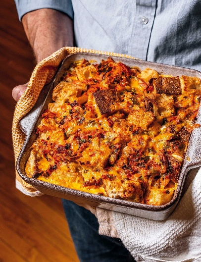 A person holding a casserole dish full of bread pudding with melted cheese and tomatoes