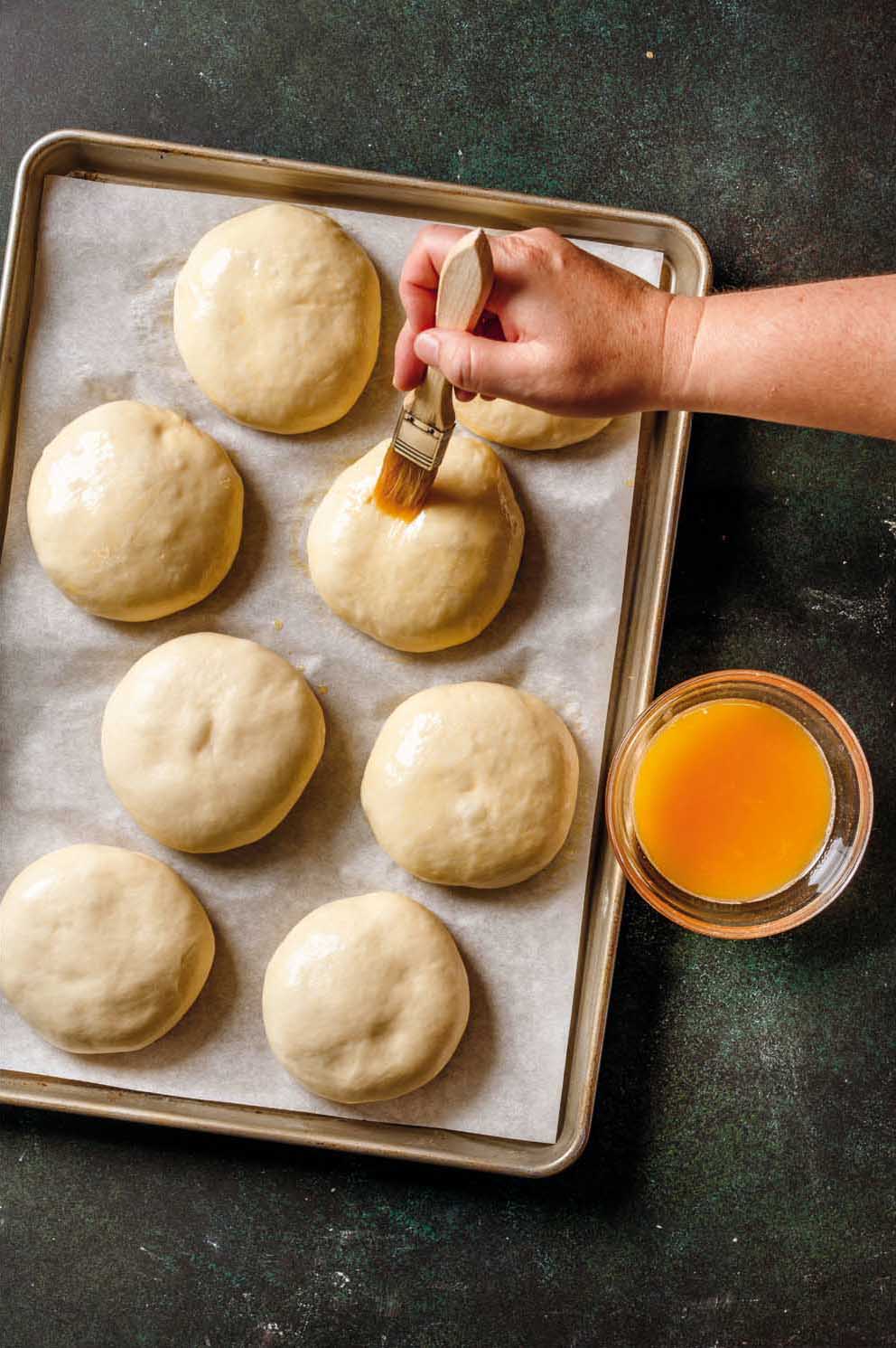 Overhead view of a baking sheet lined with parchment paper holding eight round dough balls. A hand uses a pastry brush to coat one dough ball with an egg wash, while a small glass bowl of orange-yellow egg mixture sits beside the tray on a dark countertop.